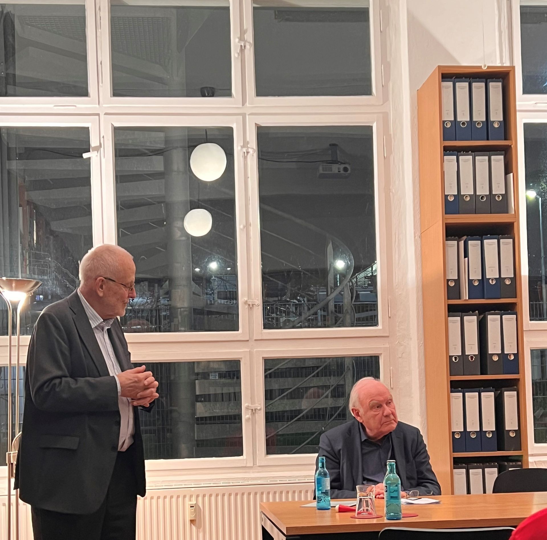 Zwei Männer sitzen an einem Tisch und unterhalten sich bei einer Buchveranstaltung. Große Fenster, Aktenschrank.