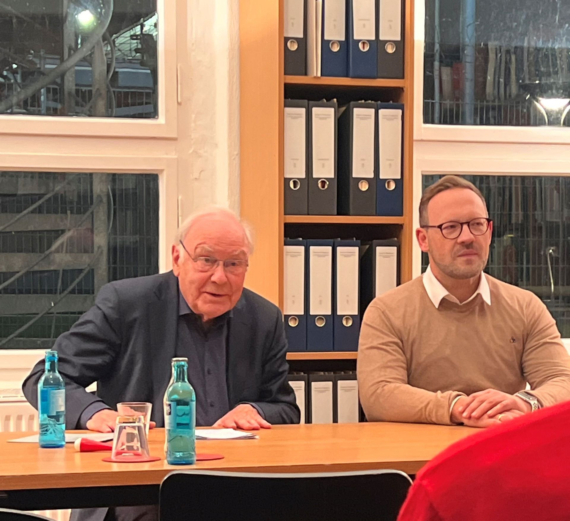 Zwei Männer sitzen an einem Tisch, einer älter, einer jünger; hinter ihnen stehen Wasserflaschen und Ordner auf einem Regal.
