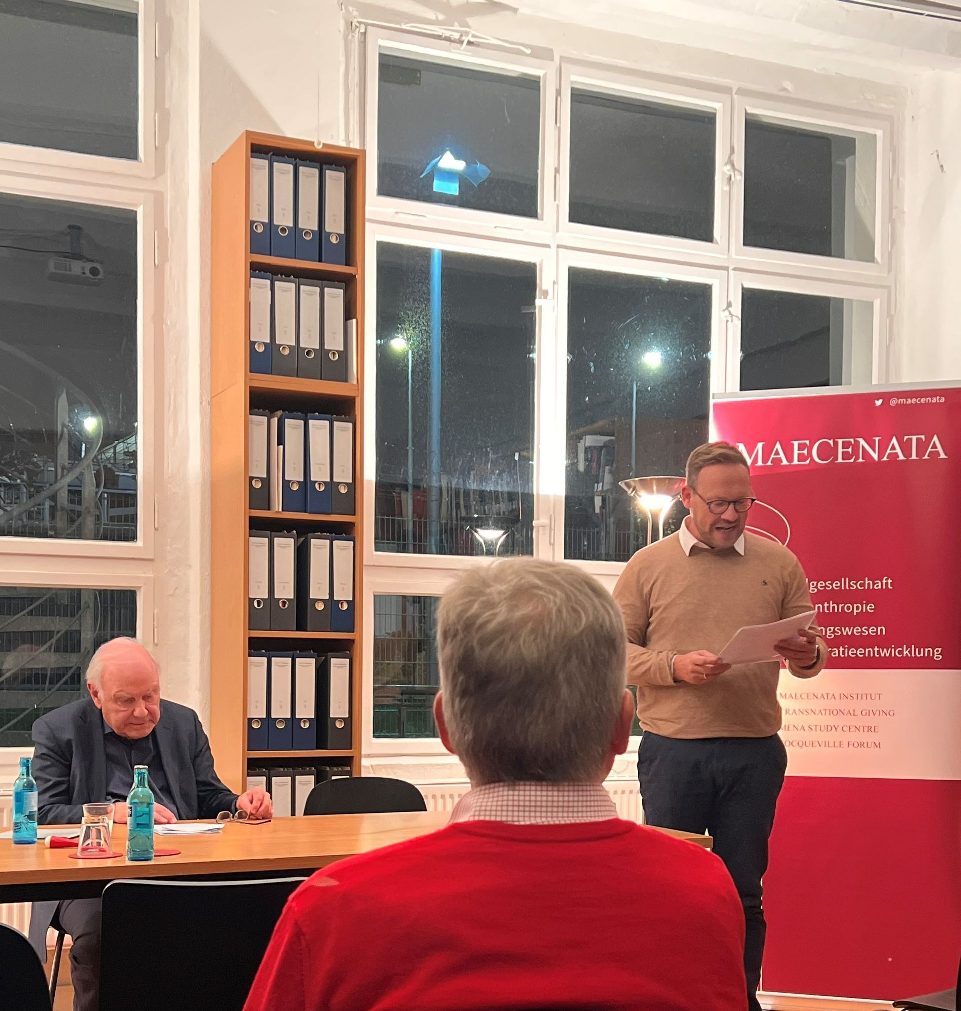 Ein Mann spricht an einem Rednerpult, zwei weitere Personen sitzen an einem Tisch vor einem Banner. Die Szene spielt sich in einem Raum ab.