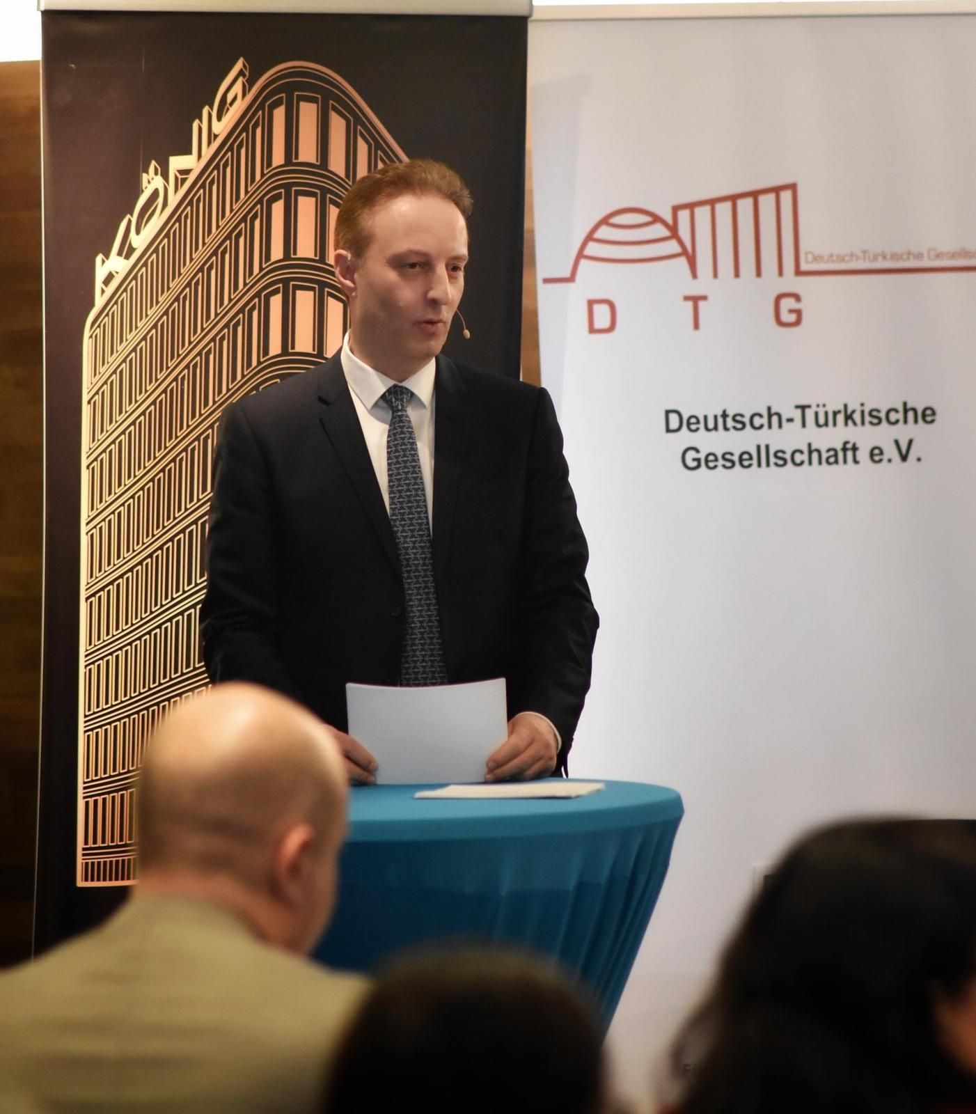 Man speaking at a podium with a Deutsch-Türkische Gesellschaft e.V. banner in the background.