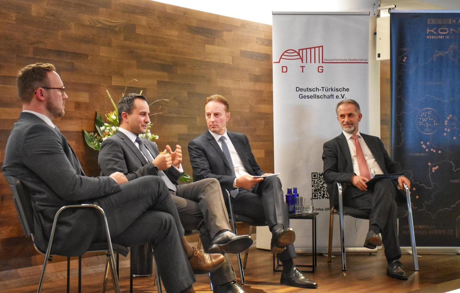 Vier Männer in Anzügen sitzen und nehmen an einer Podiumsdiskussion teil. Sie befinden sich in einem Innenraum vor einer Kulisse mit Schildern.