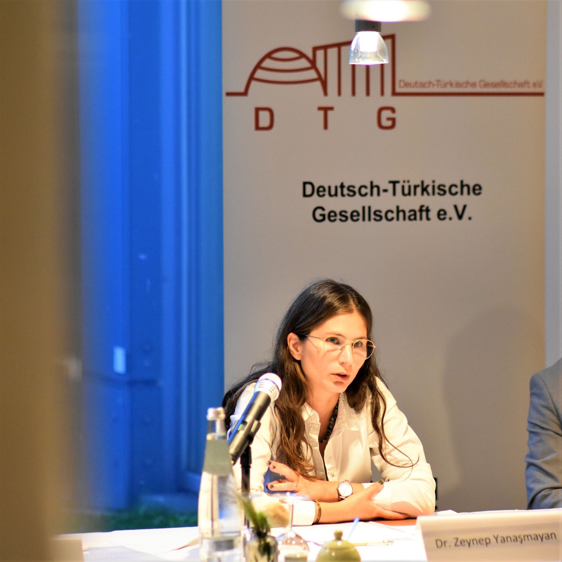 Eine Frau mit einem Mikrofon sitzt an einem Tisch vor einem Banner der Deutsch-Türkischen Gesellschaft.