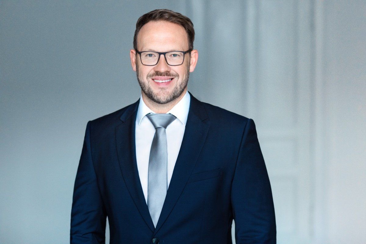 Ein Mann im Anzug mit Brille lächelt vor einem hellgrauen Hintergrund.