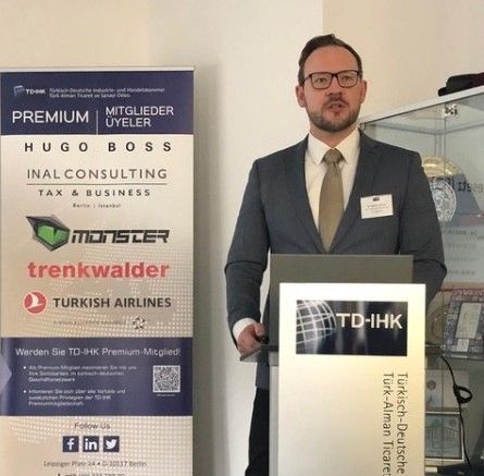 Ein Mann spricht an einem Rednerpult mit Logos, darunter „TD-IHK“ und Sponsoren wie „Hugo Boss“ und „Turkish Airlines“.