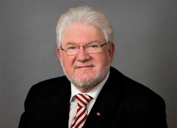 Mann mit Brille und Anzug, lächelnd, mit weißem Bart und weißem Haar; vor grauem Hintergrund.