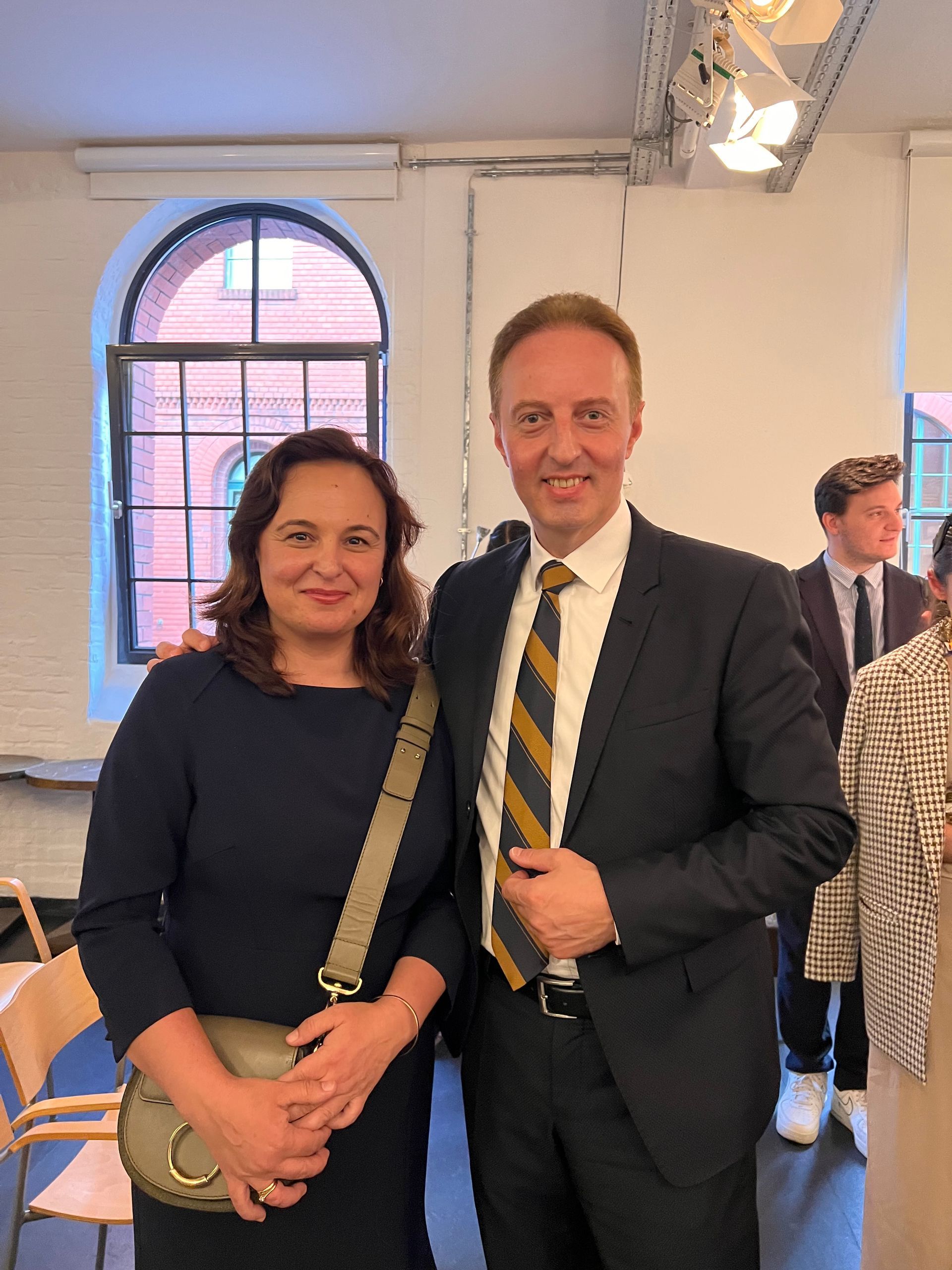 Eine Frau und ein Mann posieren in einem Innenraum. Der Mann trägt Anzug und Krawatte. Die Frau trägt ein Kleid und hat eine Handtasche.