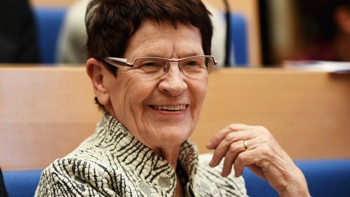 Frau mit Brille lächelt, trägt einen gemusterten Blazer, in einem Innenraum.