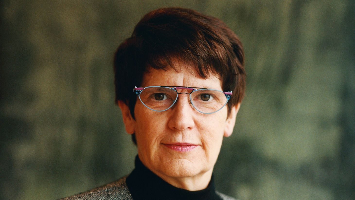 Frau mit kurzen dunklen Haaren und Brille; lächelt, trägt einen schwarzen Rollkragenpullover und eine grau gemusterte Jacke.