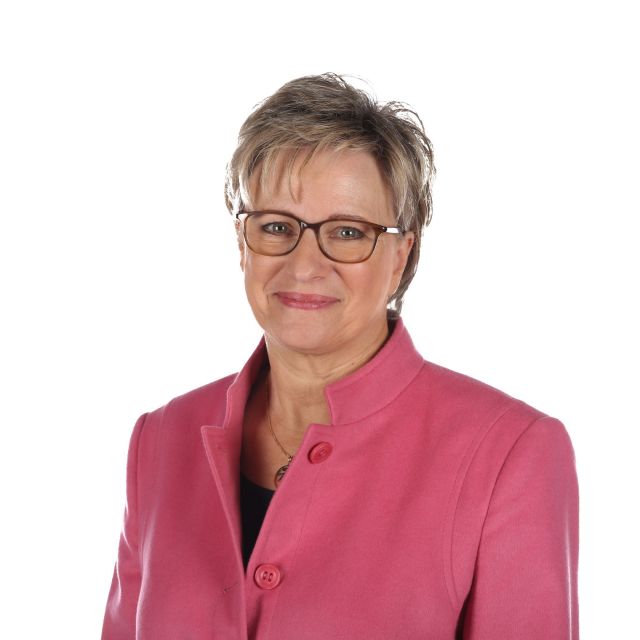Eine Frau mit Brille und rosa Jacke lächelt vor einem weißen Hintergrund in die Kamera.