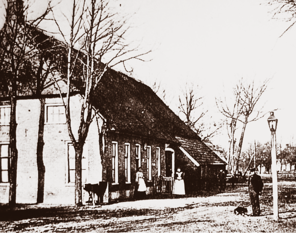 Een historische zwart-witfoto van een huis met een rieten dak, bomen en mensen die buiten staan.