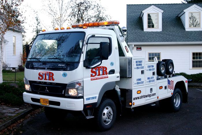 White tow truck with orange lights and red lettering parked in a driveway beside a house.