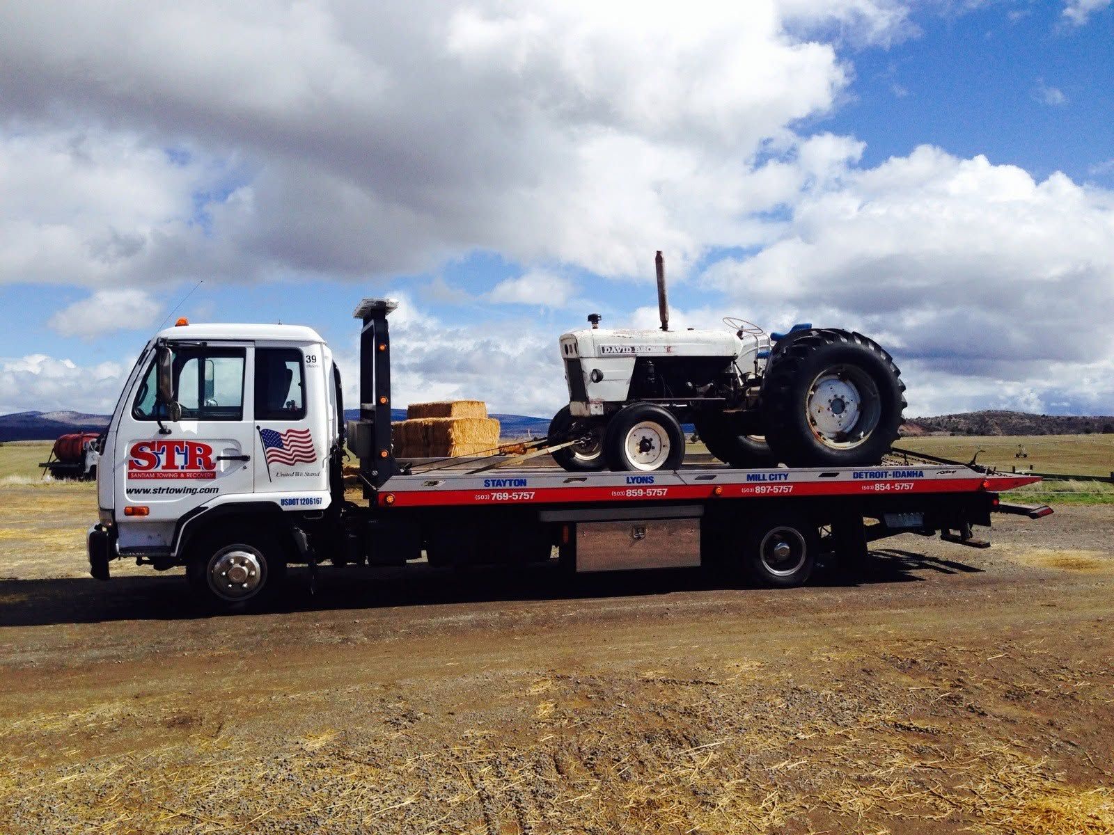 Flatbed tow truck carrying a white tractor in a gravel lot under a cloudy sky