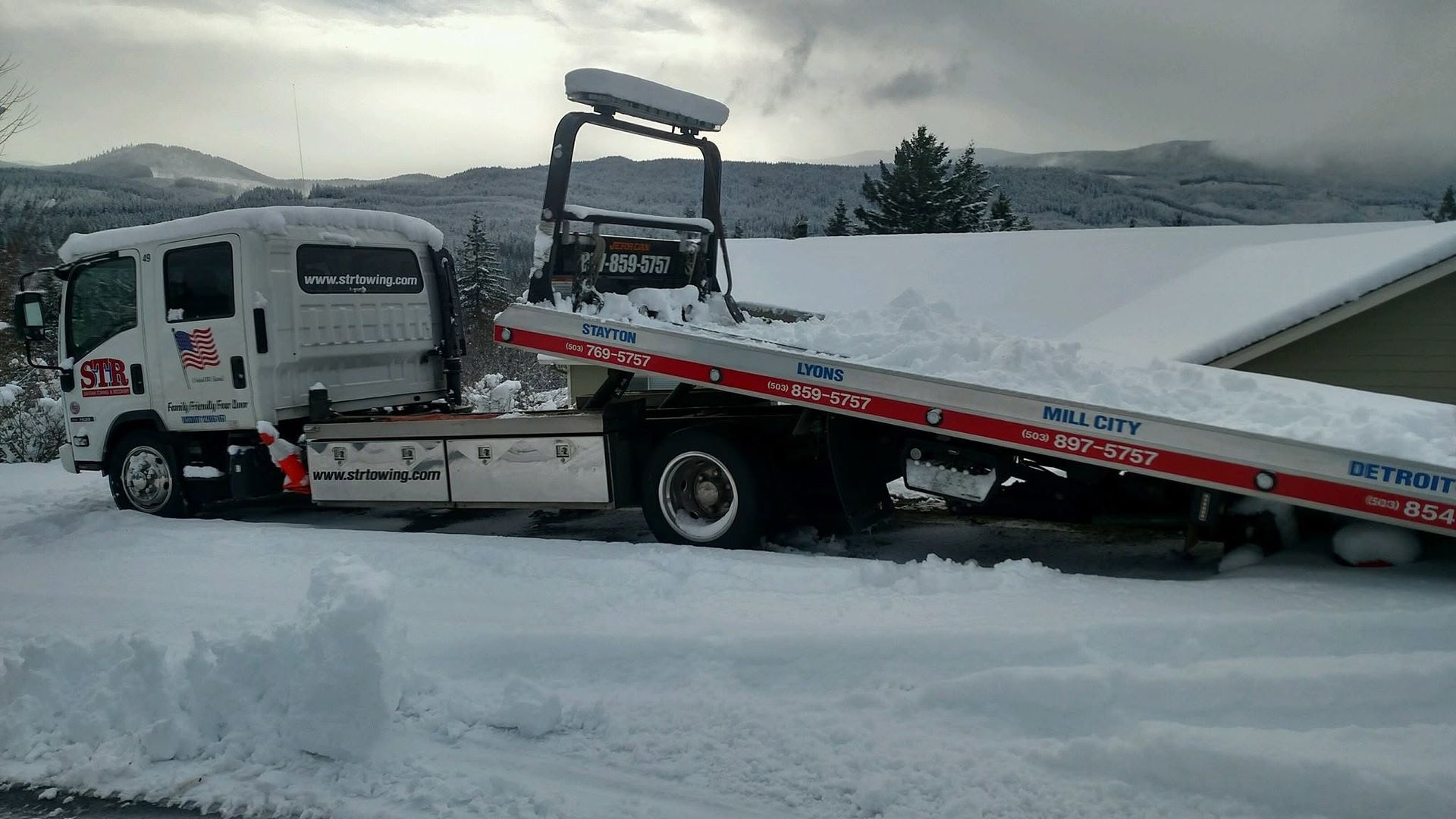 Tow truck hauling a snow-covered flatbed on a snowy road with mountains in the background