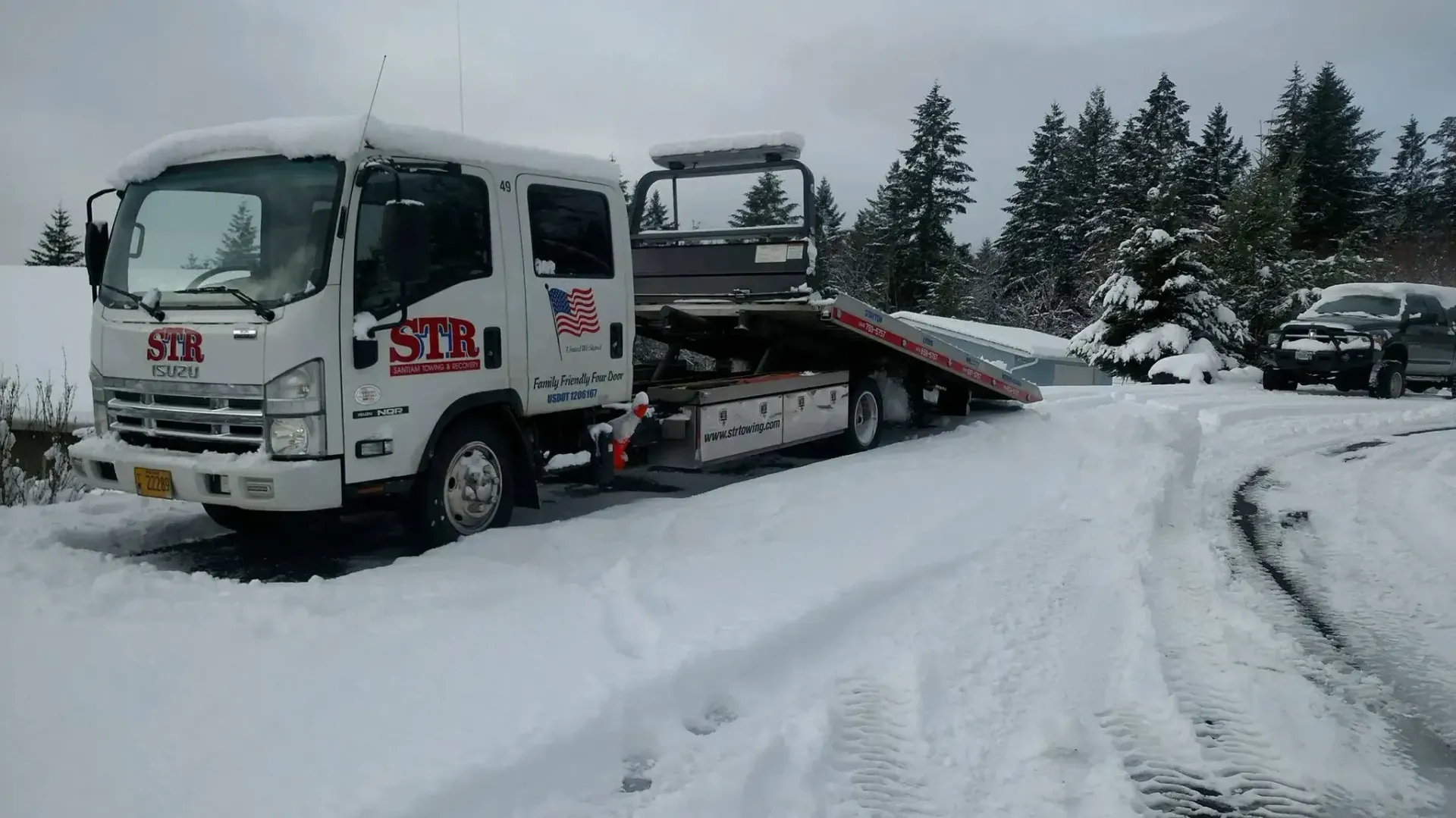 Tow truck pulling a white car in a snowy roadside scene with trees and heavy snow.