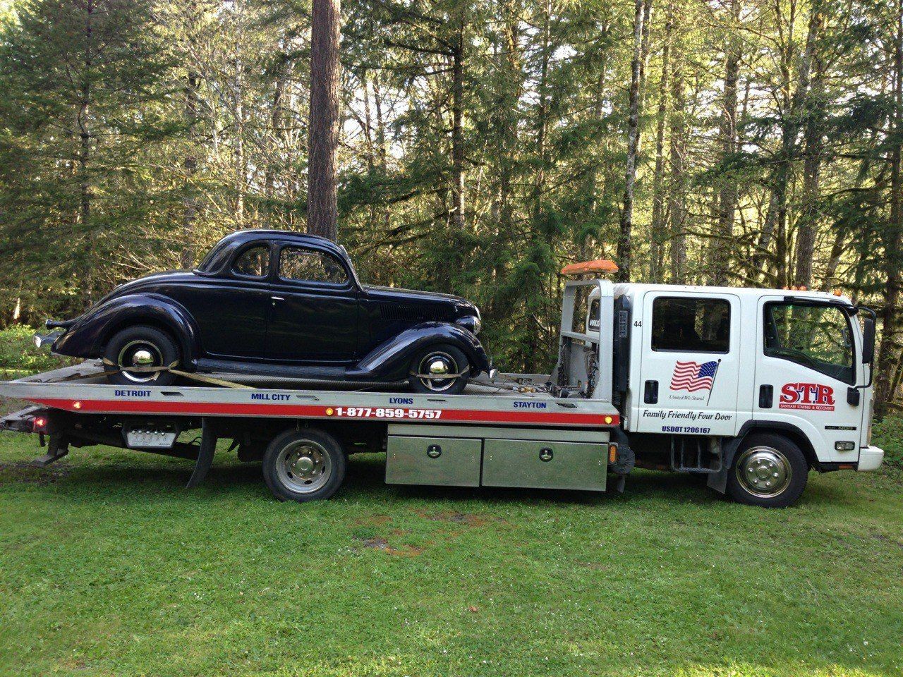 Black vintage car on a white flatbed tow truck in a grassy field with trees