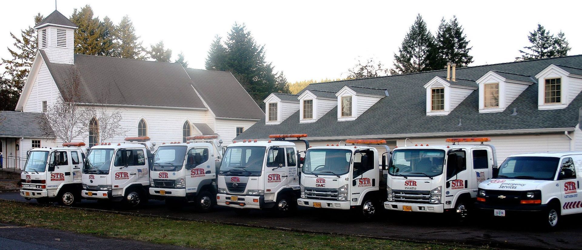 Line of white utility vans parked in front of a white church and trees