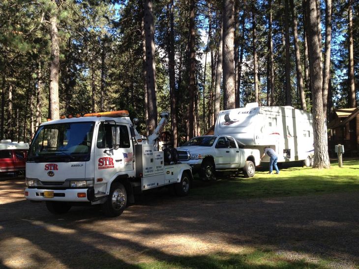 Tow truck and pickup with camper parked in a wooded campsite, with a person by the camper