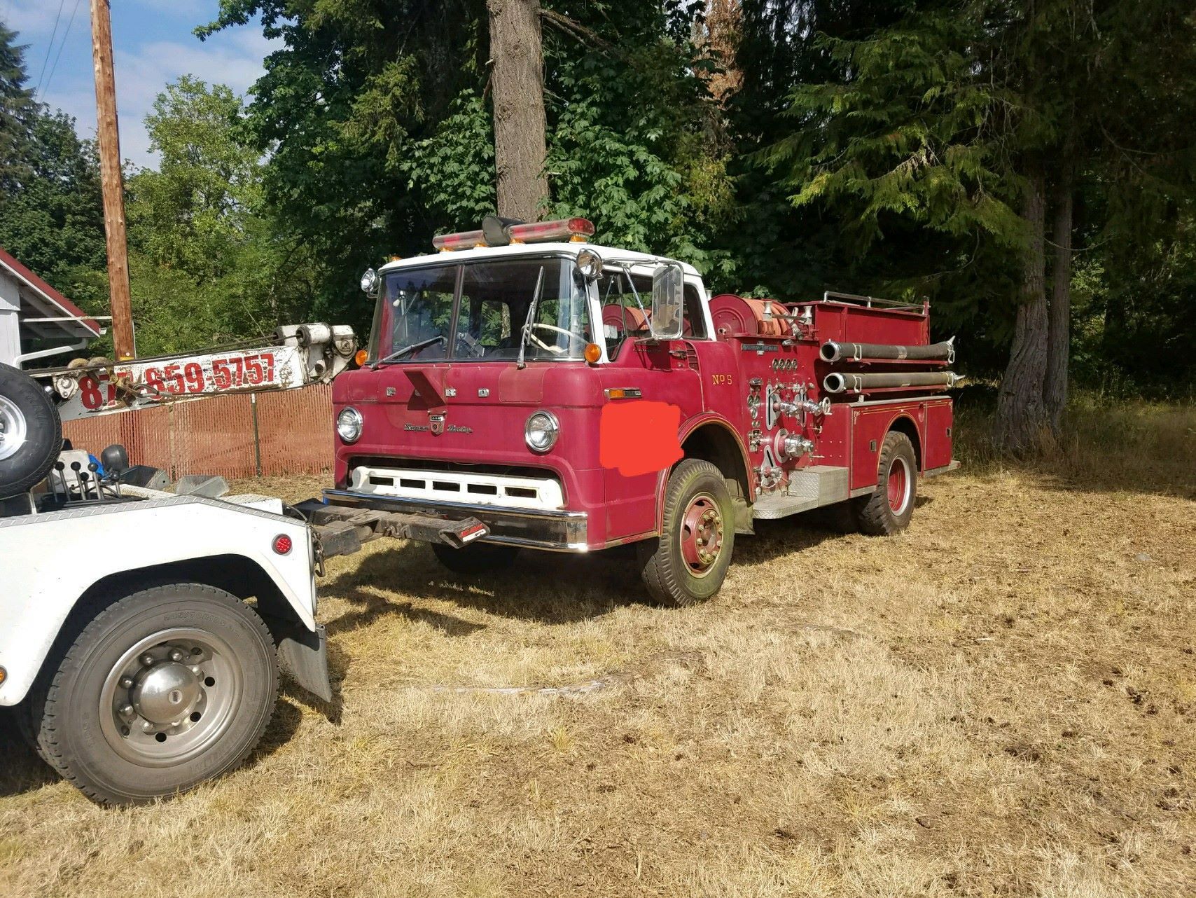 Red vintage fire truck parked on dry grass near trees, with a white vehicle partly visible in front.
