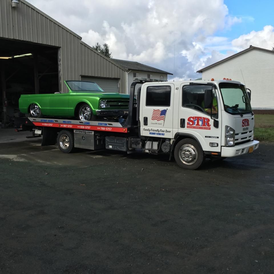 White flatbed tow truck carrying a bright green classic car outside a garage