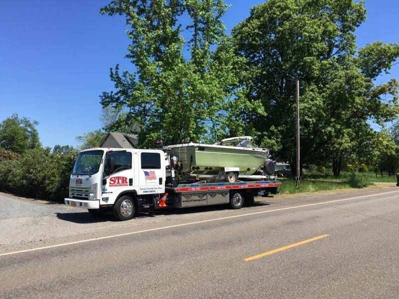 White tow truck carrying a green boat on a flatbed along a tree-lined road