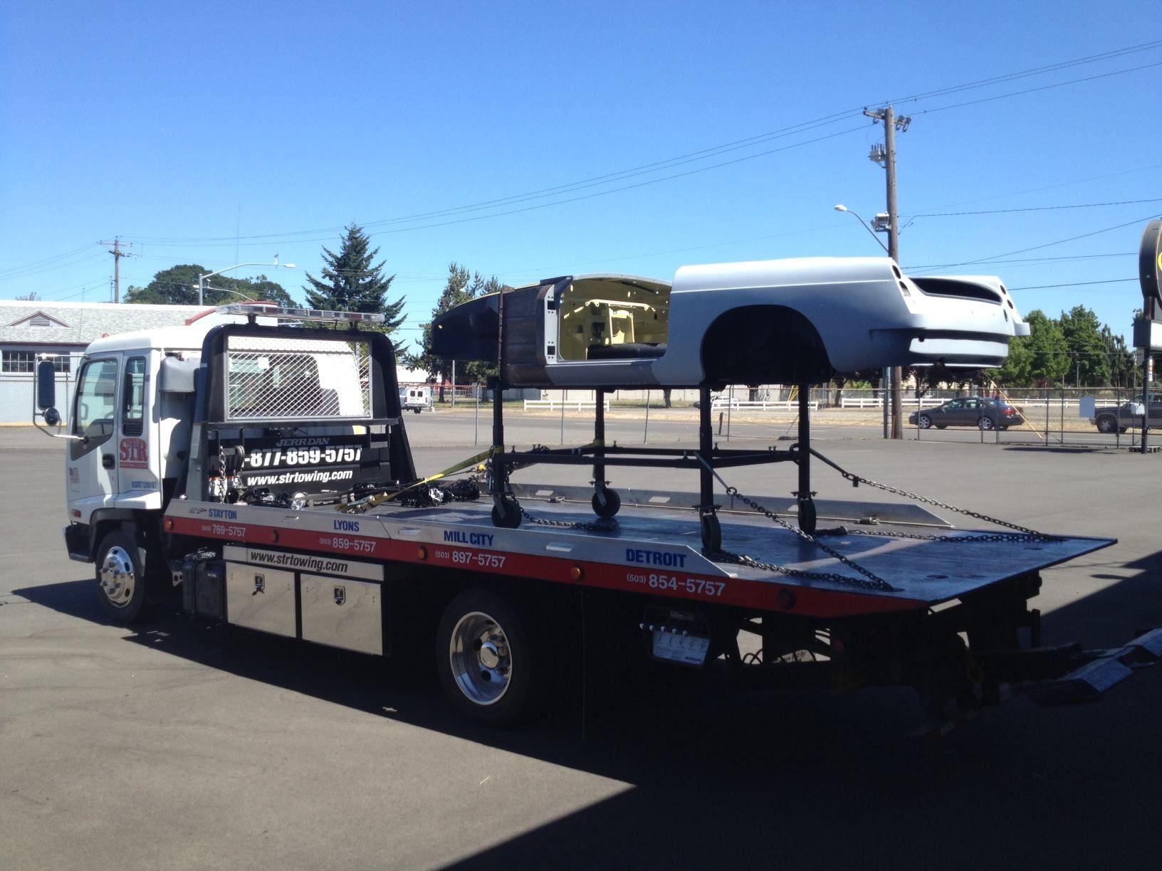 Tow truck carrying a white camper shell on a flatbed in a parking lot under a blue sky