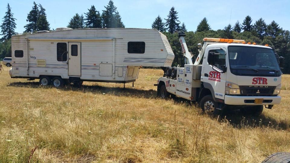 Tow truck towing a white camper trailer in a dry grassy field with trees in the background