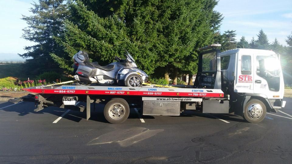 Tow truck carrying a silver scooter on a flatbed in a parking lot, with trees in the background