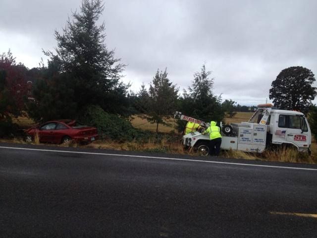 Tow truck and two workers beside a red car on a wet roadside under cloudy skies
