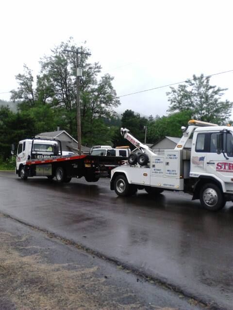 Tow truck hauling a white car on a wet roadside in a rainy neighborhood