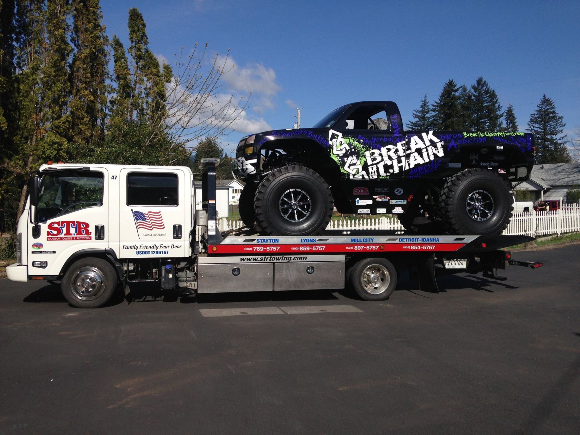 White flatbed tow truck carrying a black lifted monster truck outdoors on a sunny day