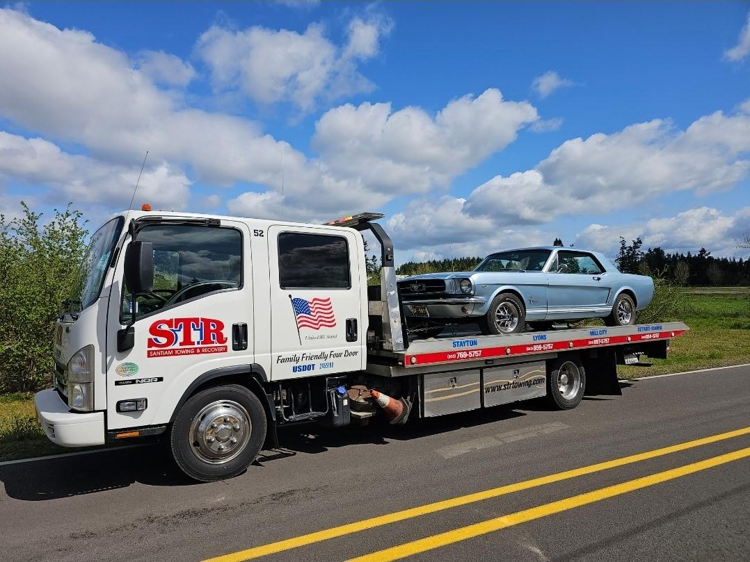 Tow truck hauling a silver car on a flatbed beside a rural road under a blue sky