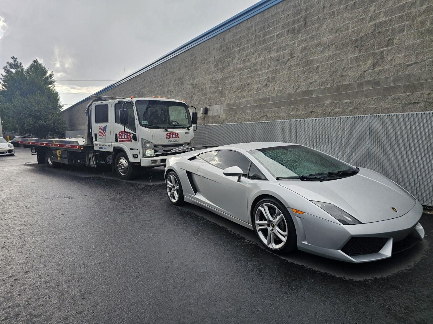 Silver sports car parked beside a tow truck on a wet road next to a large concrete wall.