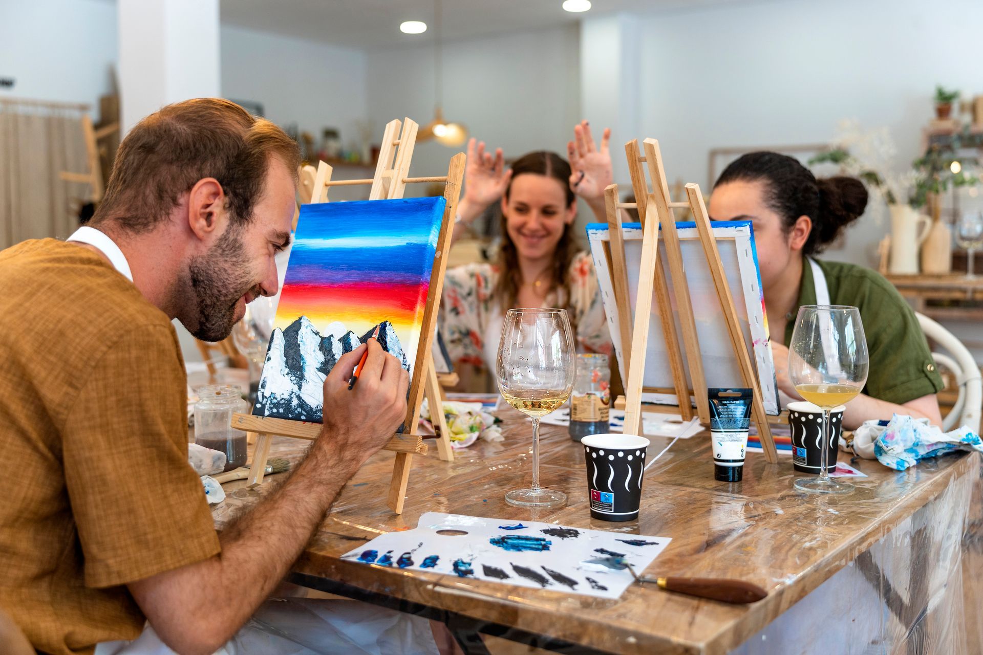 People painting at easels in an art class; colorful sunset scene, smiles, wine glasses on table at EVOQ East Cobb Marietta.