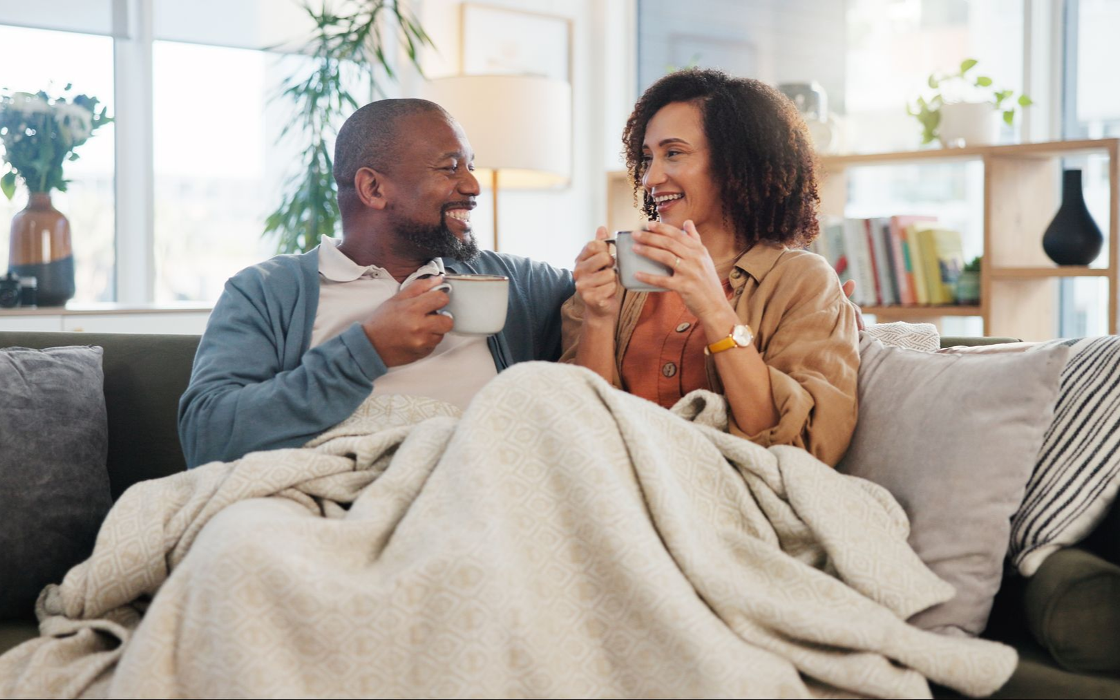 Two people smiling on a couch, wrapped in a beige blanket, holding mugs in a bright living room