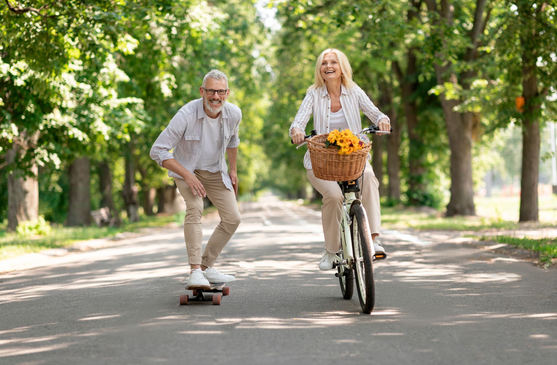 Two people ride a bicycle and skateboard on a tree-lined road in a sunny park at EVOQ East Cobb.