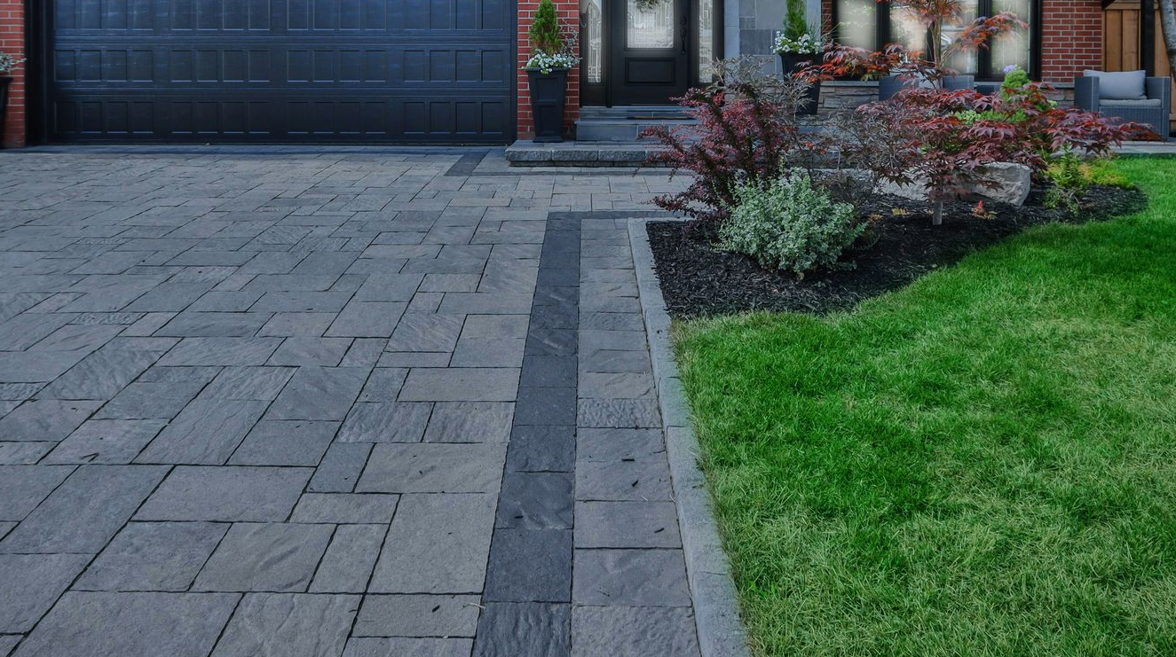 Driveway and walkway with gray pavers beside a green lawn and shrub bed in front of a garage