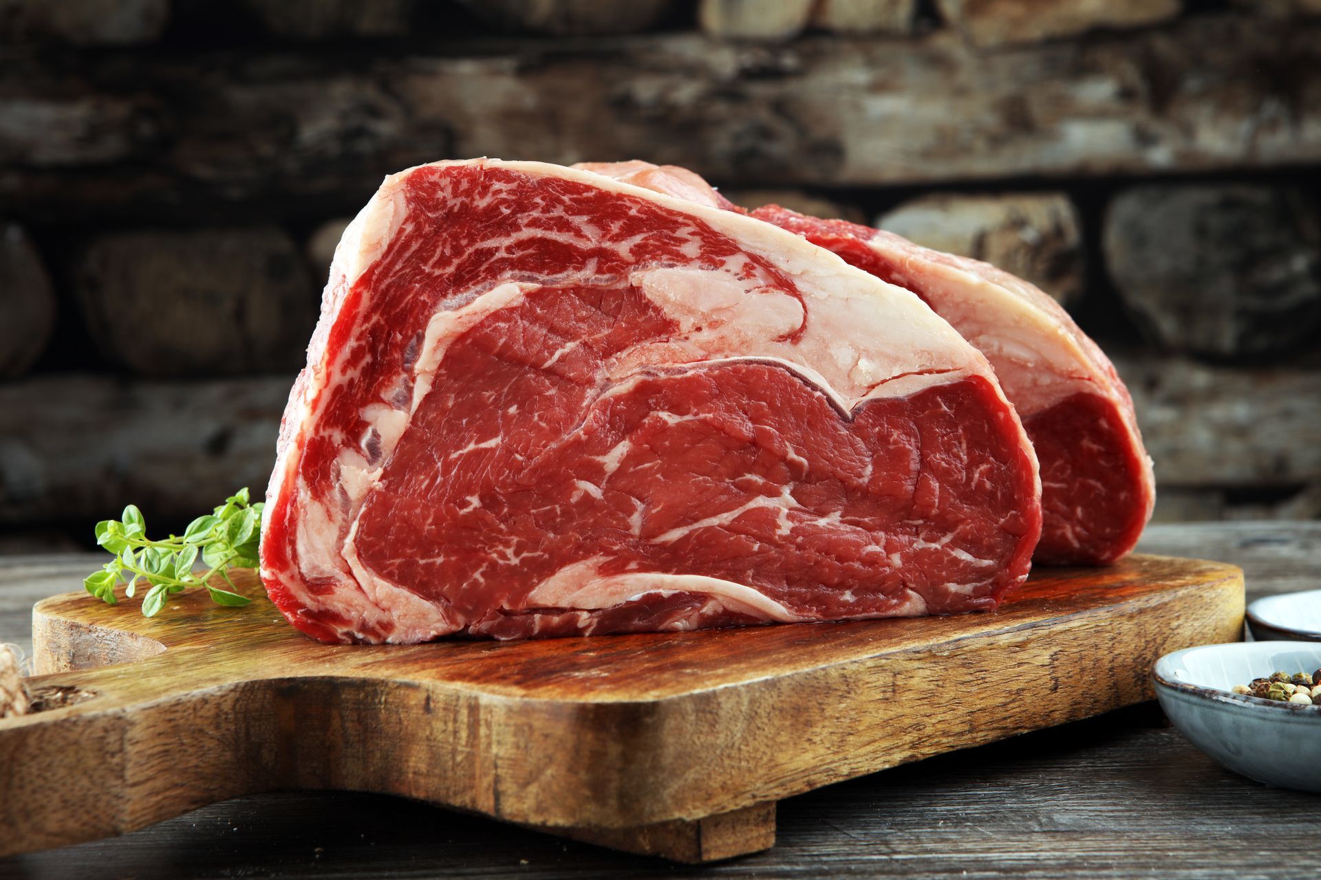 Raw marbled beef steak on a wooden cutting board with herbs and a small bowl on a rustic table