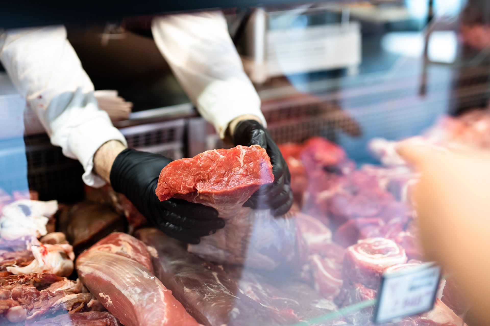 Gloved hands holding raw meat at a butcher counter with various cuts displayed in the background