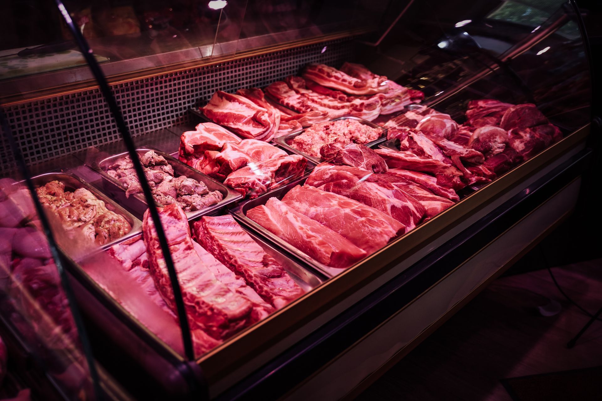 Butcher display case filled with assorted raw meats under pink lighting