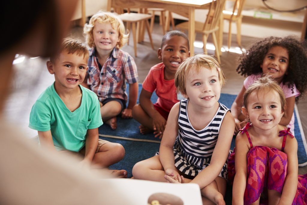 A group of children are sitting on the floor listening to a teacher read a book.