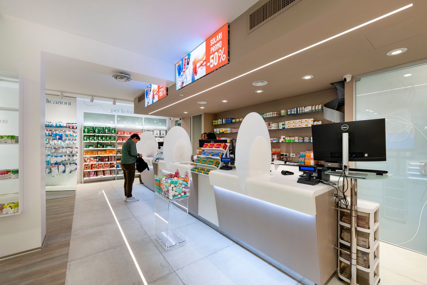 Interno moderno di una farmacia con banconi bianchi, scaffali pieni di prodotti e un cliente in fondo alla stanza.