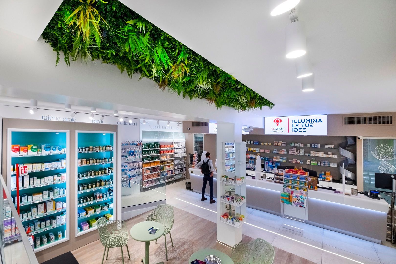Interno luminoso della farmacia con scaffali bianchi, una composizione di piante verdi appese e un membro dello staff vicino al bancone.