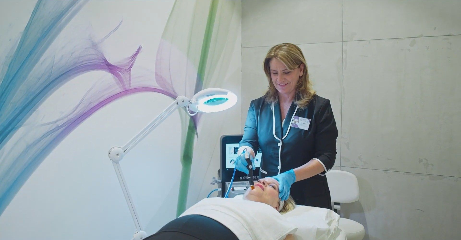 Un dentista in camice scuro utilizza uno strumento odontoiatrico su un paziente sotto una lampada d'ingrandimento in una clinica luminosa.