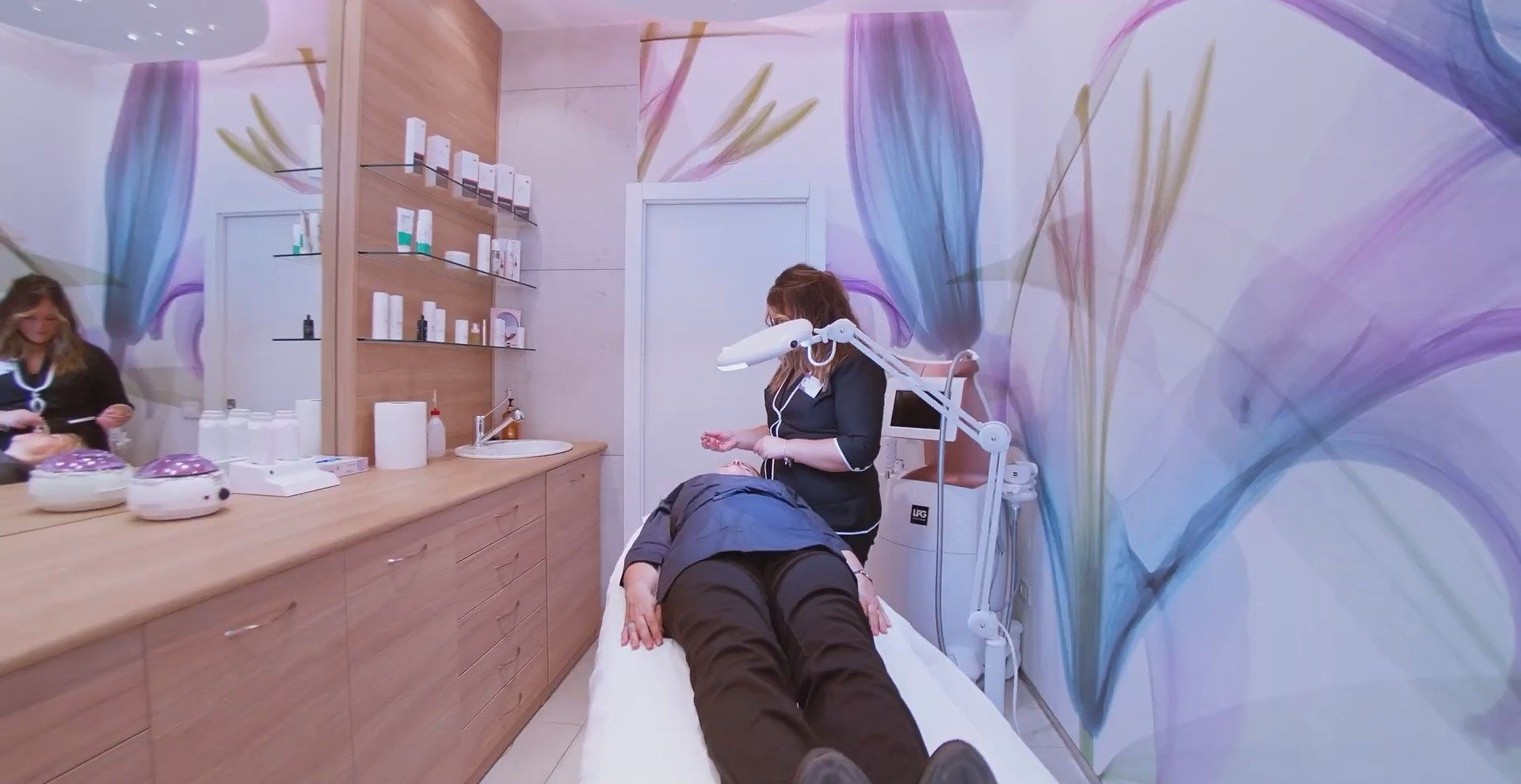 Sala trattamenti viso in un salone di bellezza, con una cliente sdraiata su un lettino e un'estetista al lavoro nelle vicinanze.