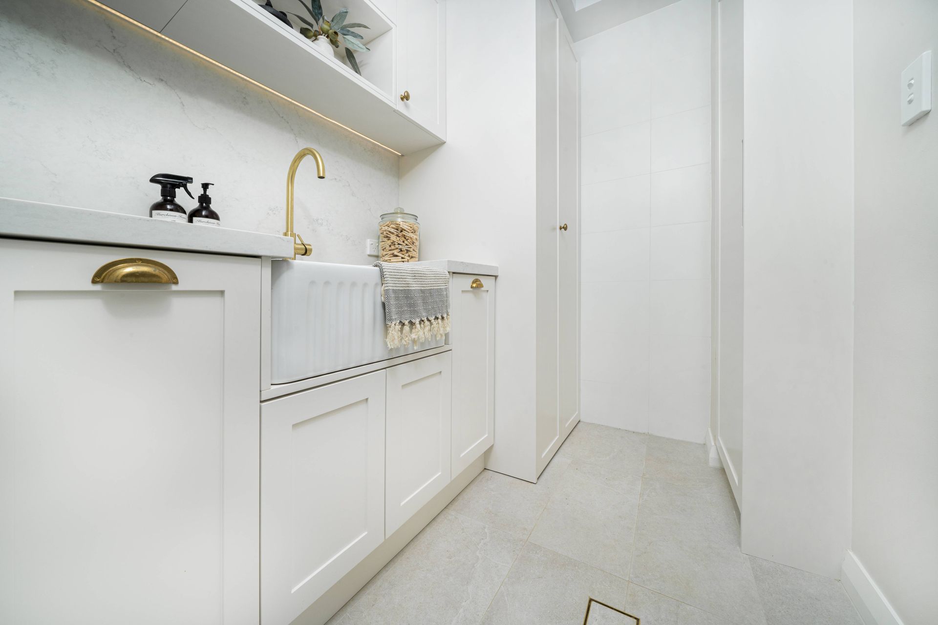 Modern white laundry room with gold tapware, built-in cabinetry, tiled floor, and hanging towel rack