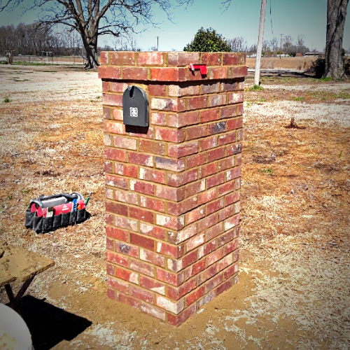 A newly built brick mailbox column standing in a yard, with a small black mailbox embedded and a red flag on top.