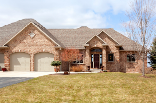 A one-story brick house with a two-car garage, brown roof, and front lawn under a cloudy blue sky.