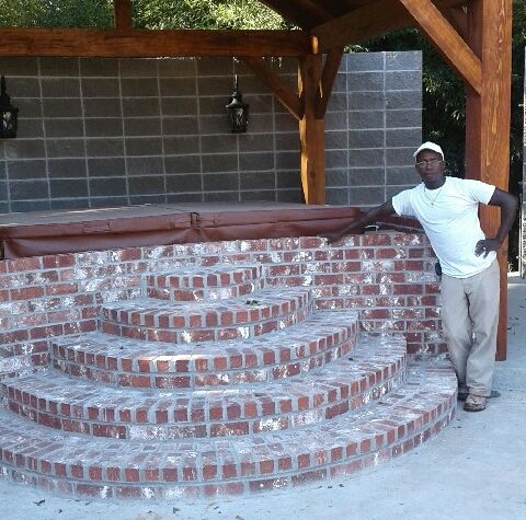 A man stands next to a freshly constructed, semi-circular set of brick steps leading up to a covered hot tub.