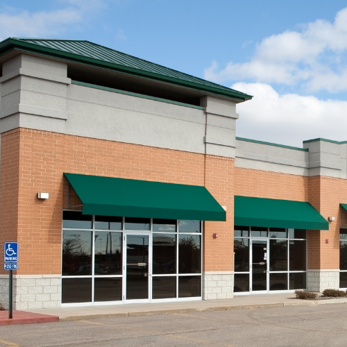 A modern brick commercial building with green awnings and glass storefronts under a blue sky.