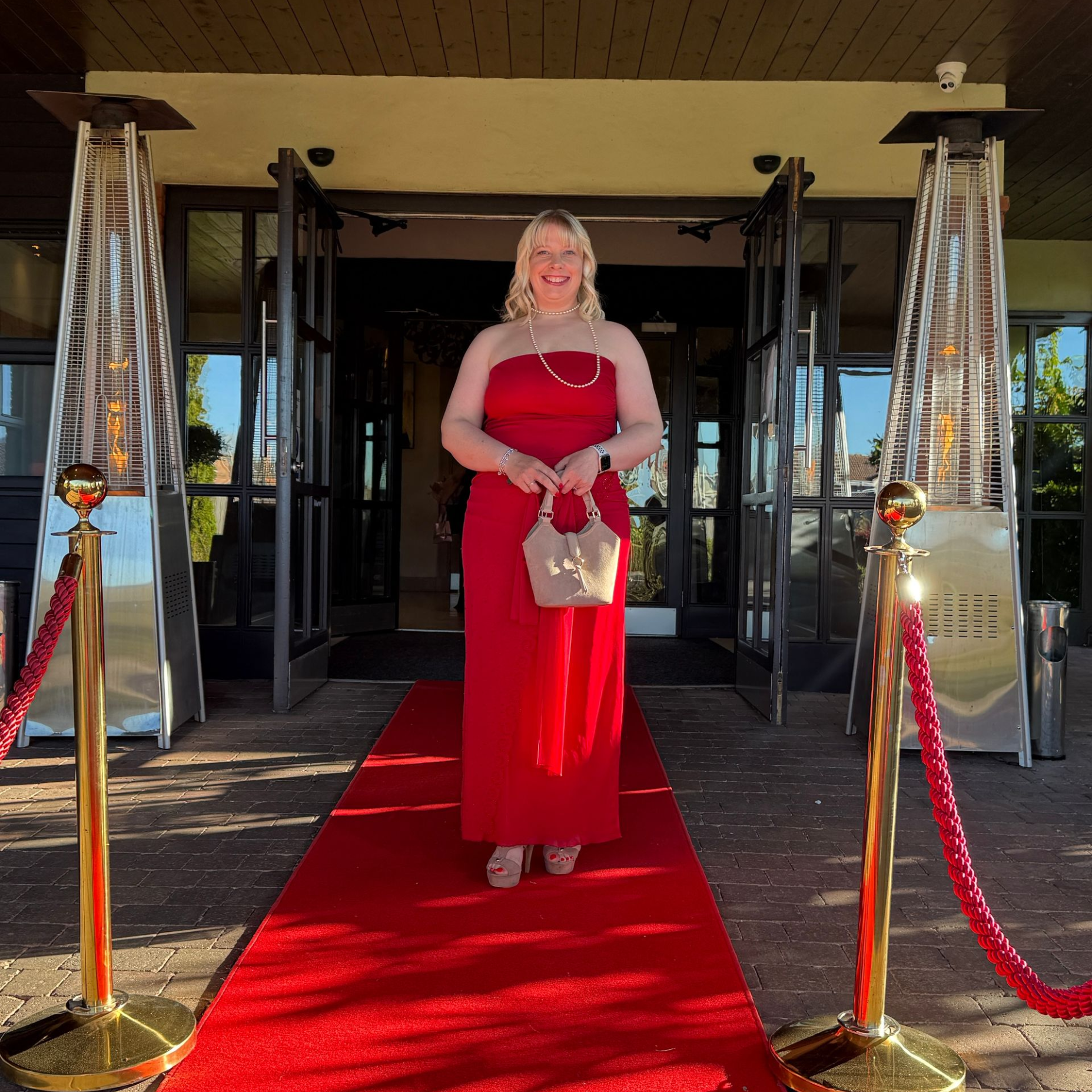 Woman in a red dress standing on a red carpet entrance, holding a small bag.